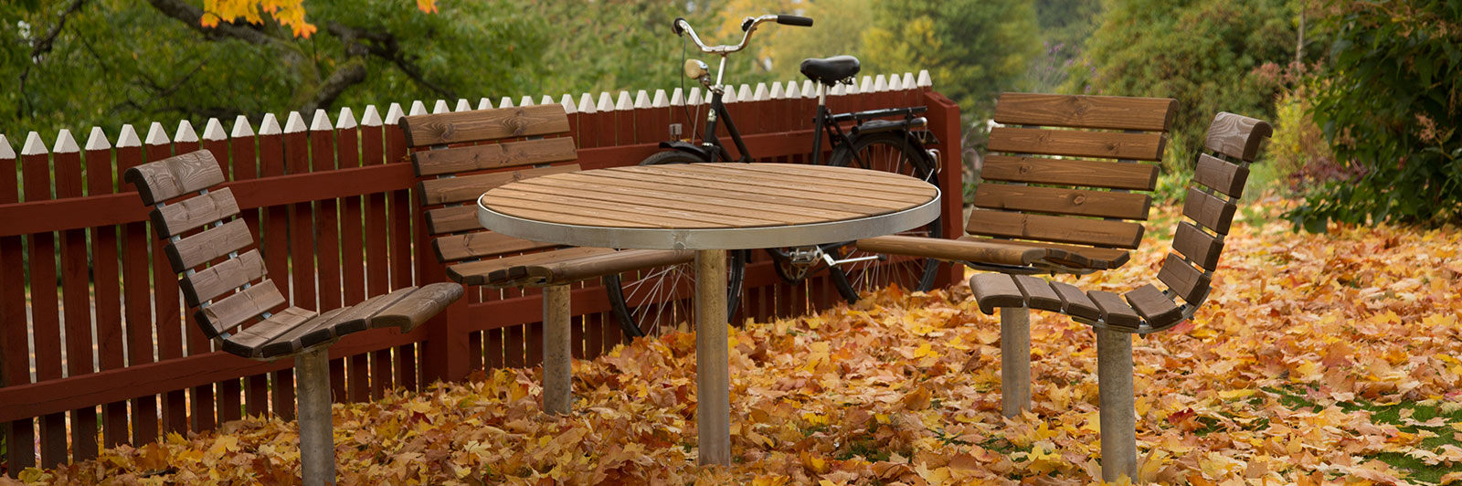 A round park table with park chairs around it nestled in a park with fallen autumn leaves all around.